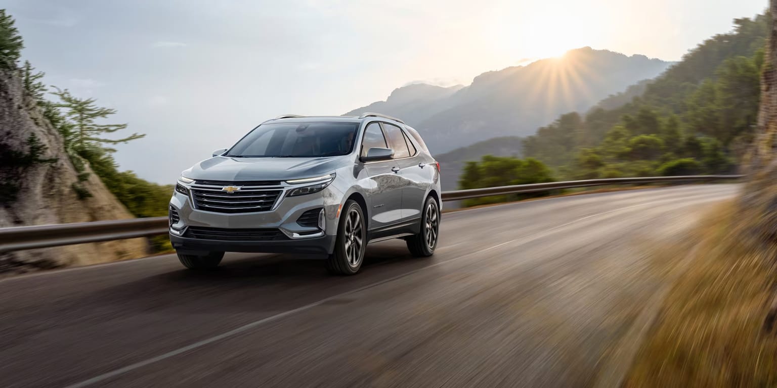 A white SUV is driving down a winding road, with a mountain in the background.