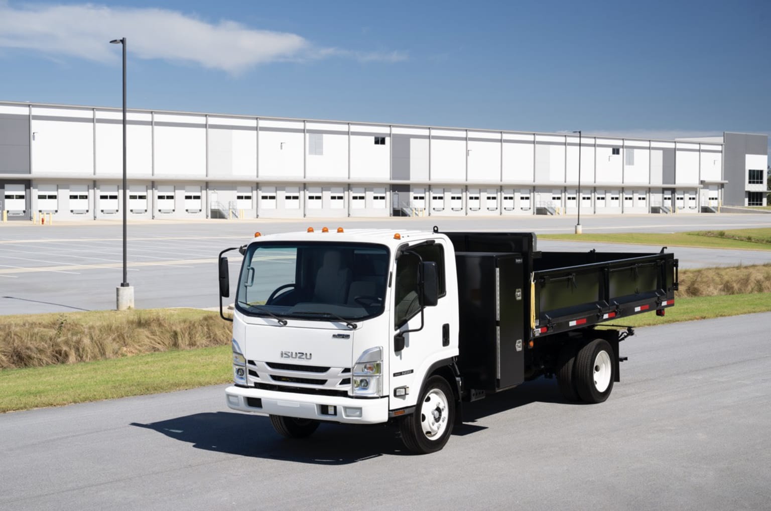 A white dump truck parked in the foreground with a large white industrial building in the background against a blue sky with some clouds.