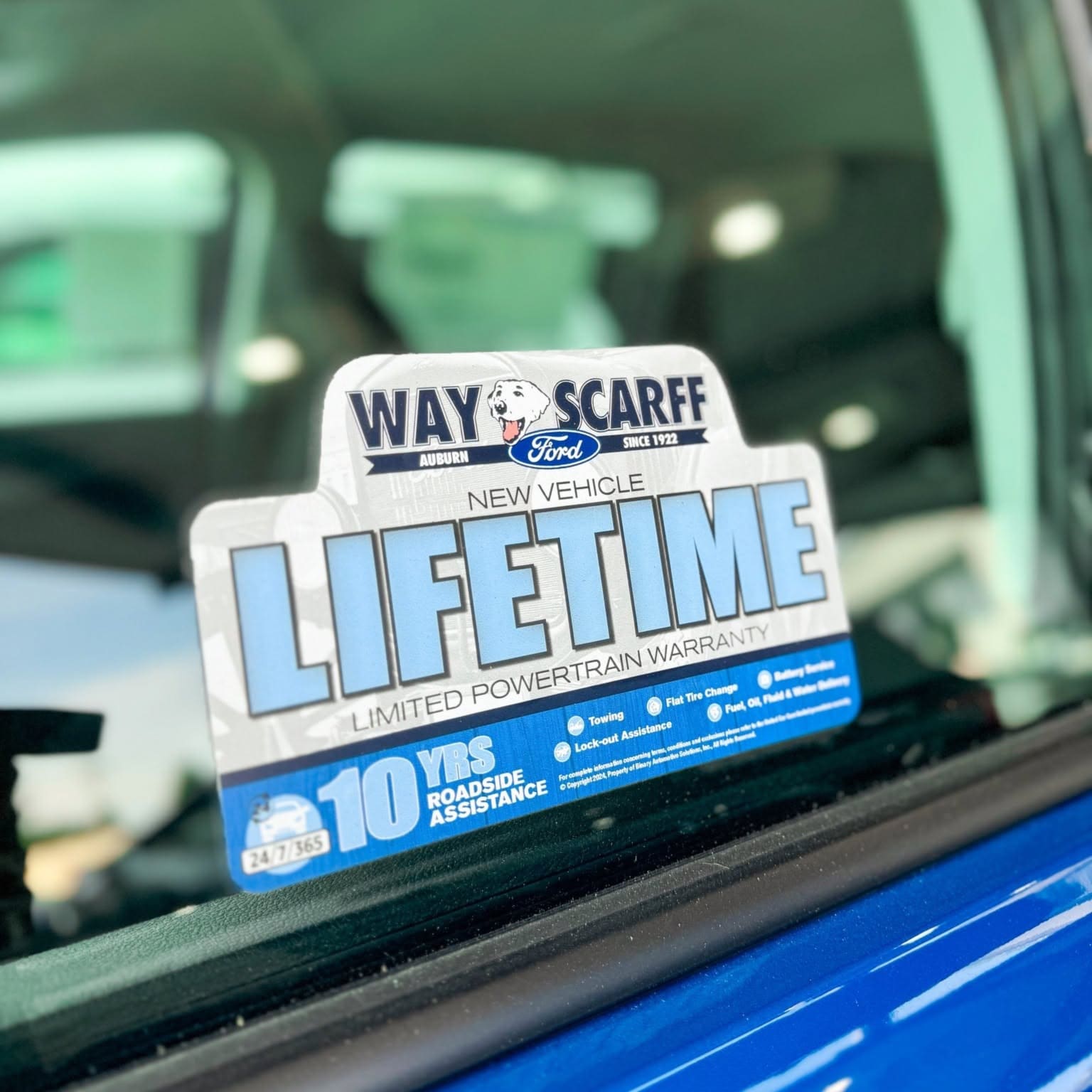 Image of a New Vehicle Lifetime Limited Powertrain Warranty sticker with 10 YRS Roadside Assistance affixed to a car window, with a blurry dealership logo and a blue car exterior visible in the background.