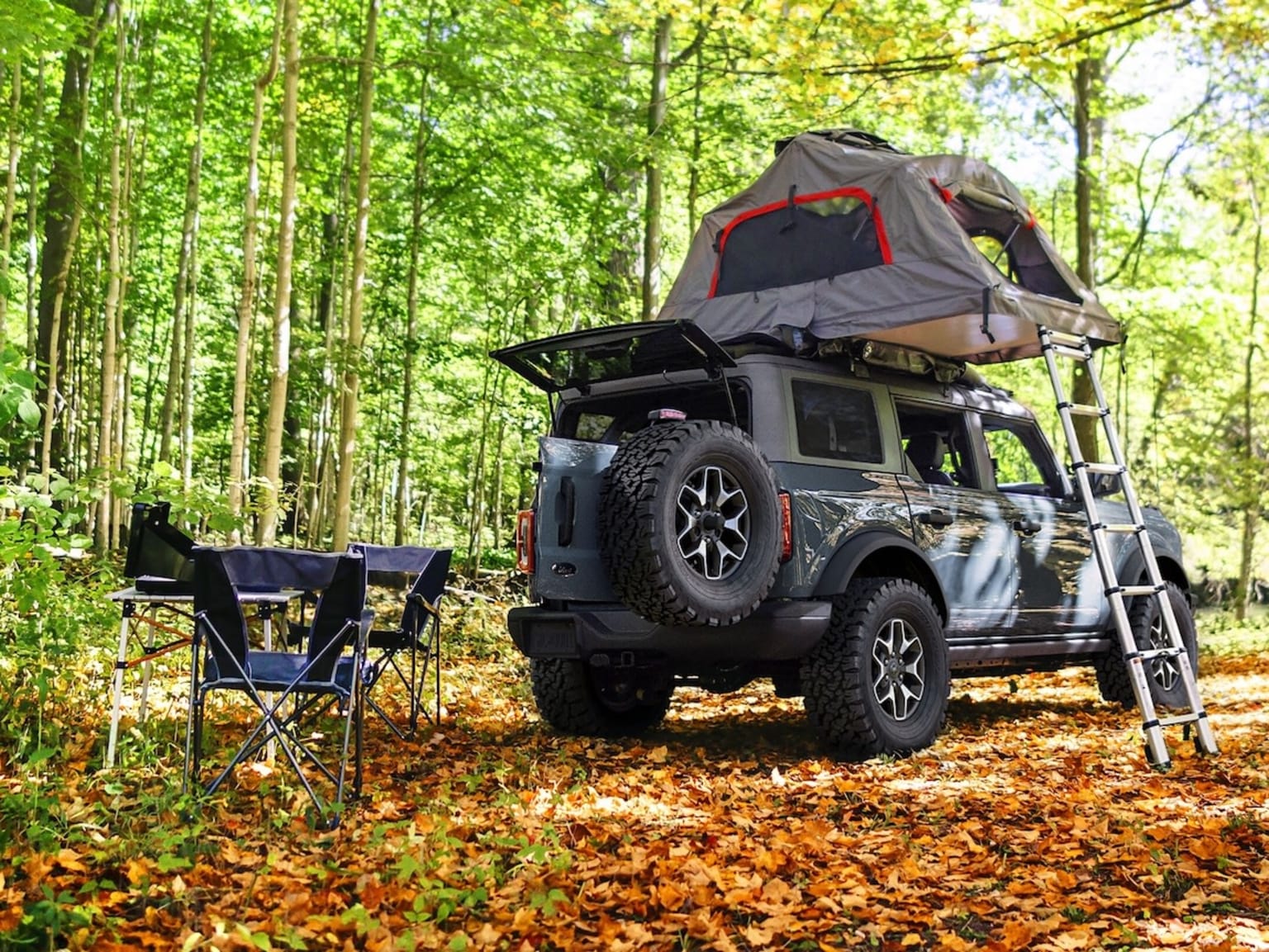 Off-road vehicle with rooftop tent parked in forest with fallen autumn leaves.