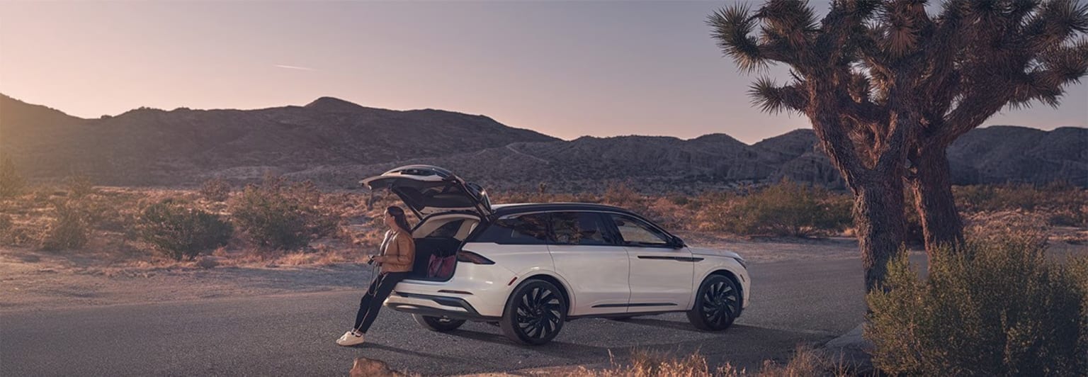 White SUV on desert road with mountains and Joshua trees in background.