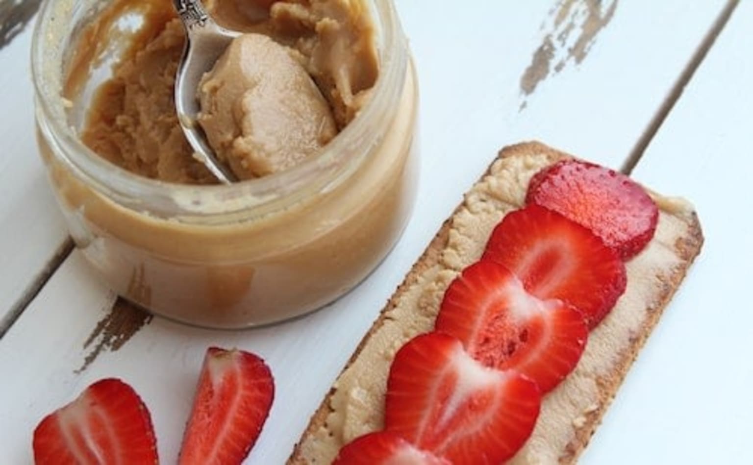 a jar of peanut butter and a sandwich with strawberries on a white wooden table with a spoon in it