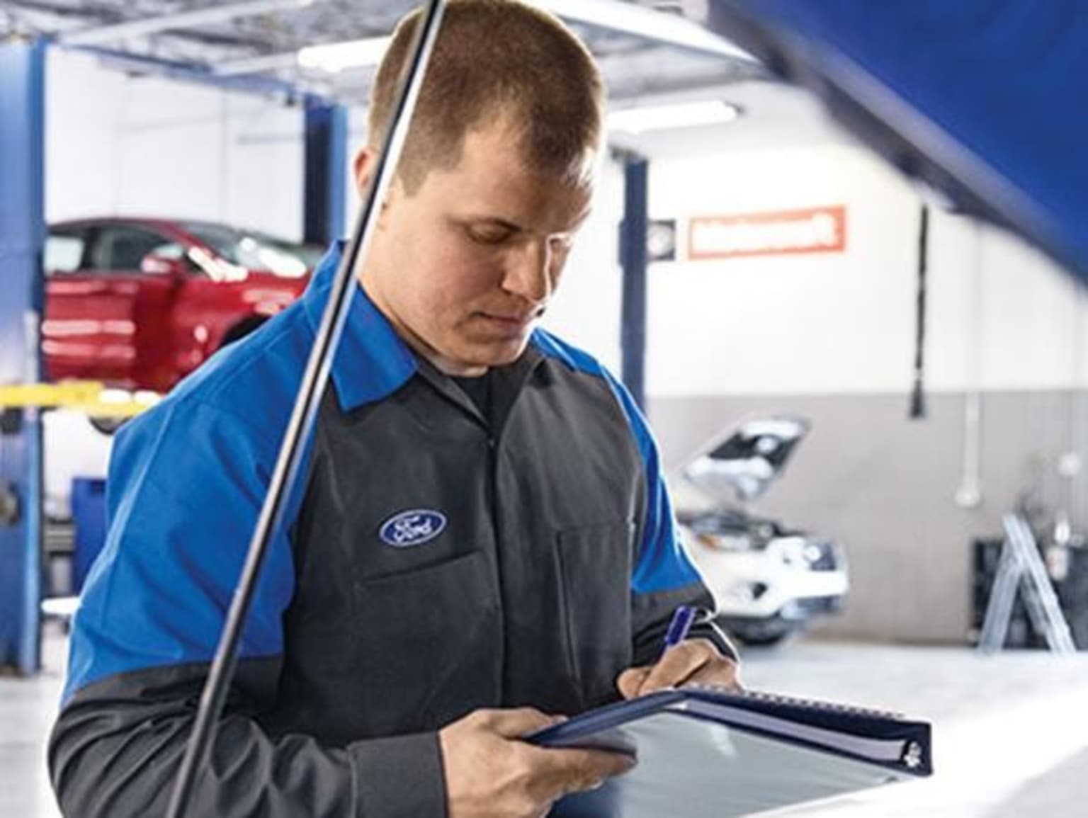 An auto mechanic in uniform is using a tablet device in a car repair shop with various tools and equipment visible in the background.
