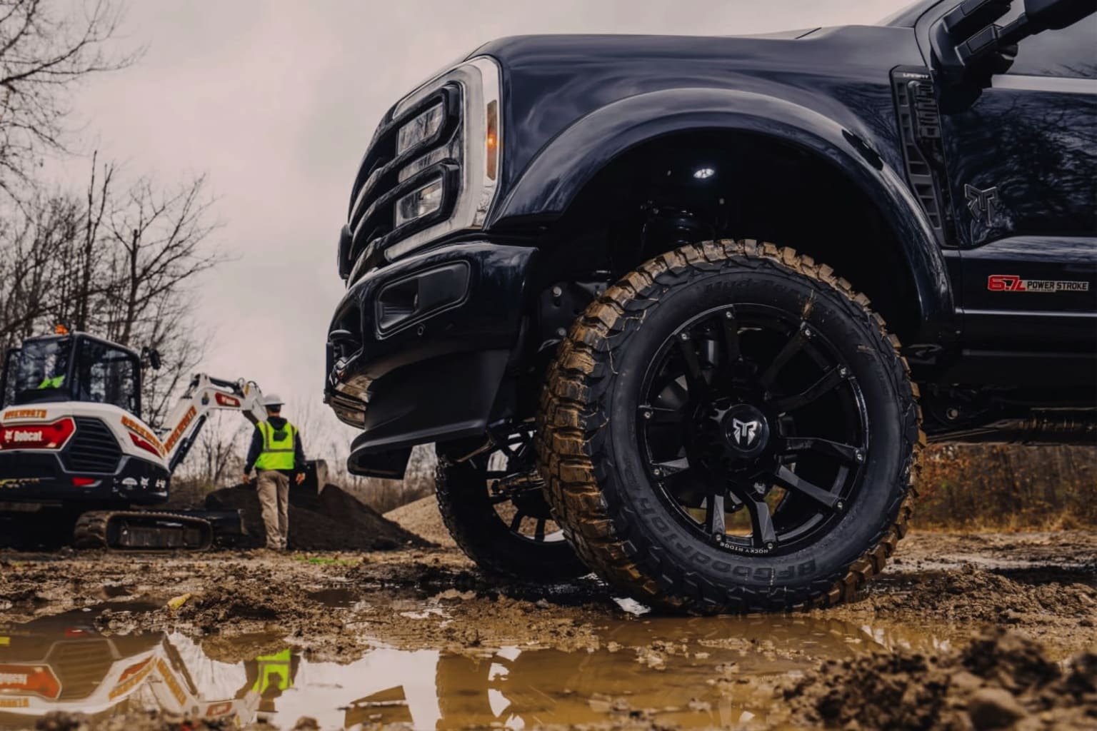 An off-road vehicle with large tires parked in a muddy, wooded area, with another similar vehicle visible in the background.