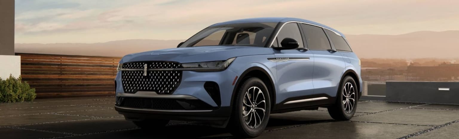 A blue SUV is parked on a paved surface in front of a wooden wall and a hazy landscape.