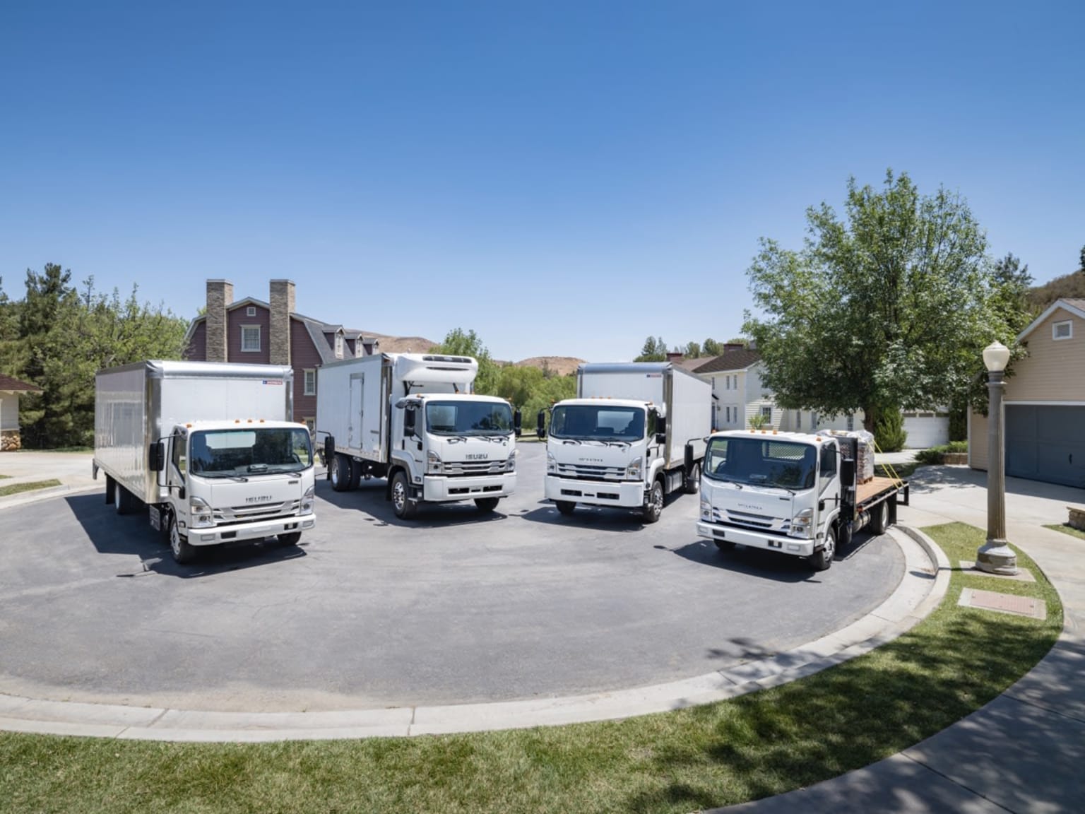 Four white delivery trucks parked in a circle on a paved area with houses and trees in the background under a clear blue sky.