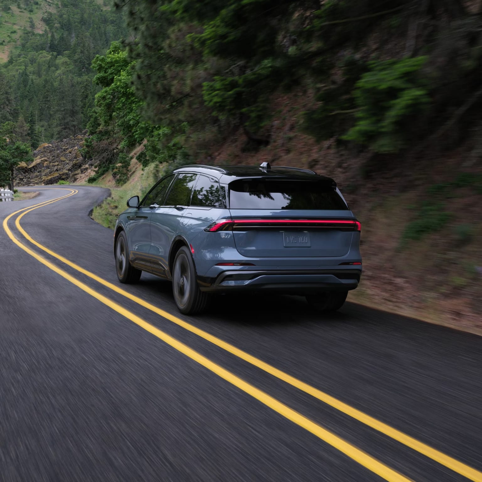 A gray SUV driving on a winding mountain road surrounded by lush green foliage and a rugged rocky landscape.