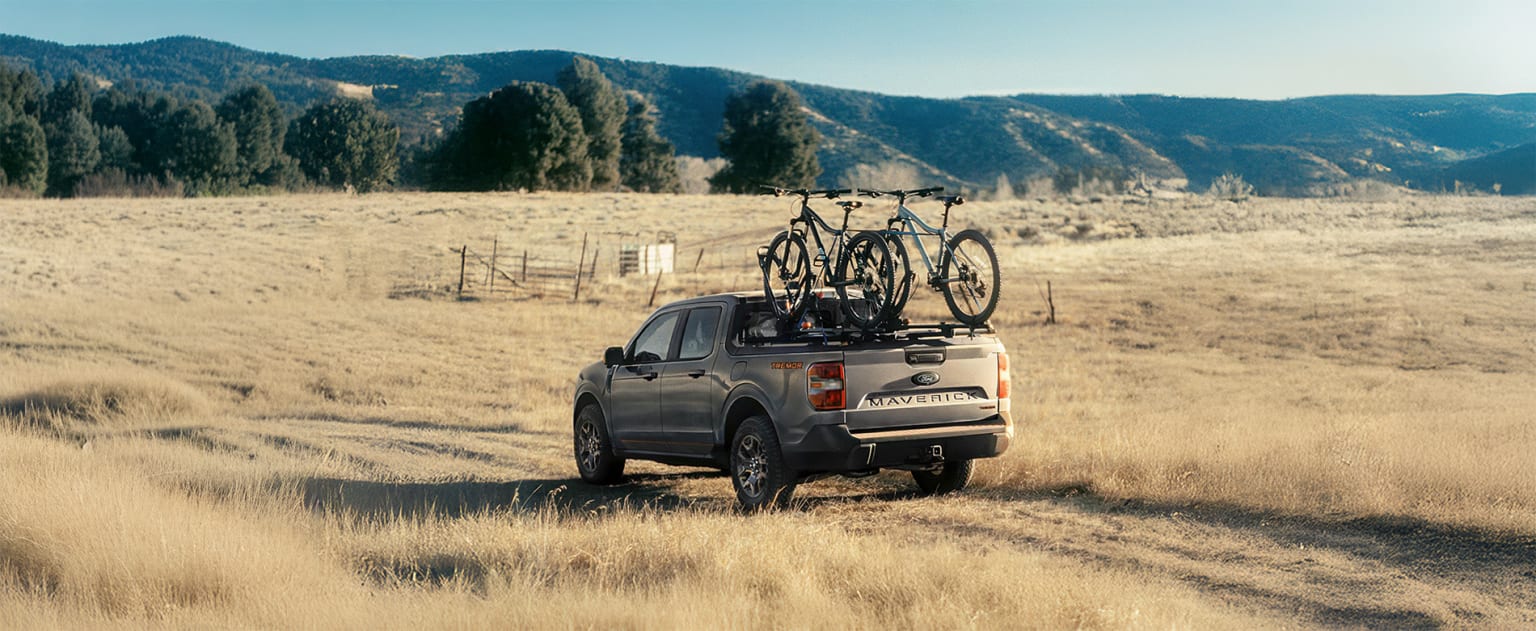 A pickup truck with a bicycle rack parked in a grassy field with mountains and trees in the background.