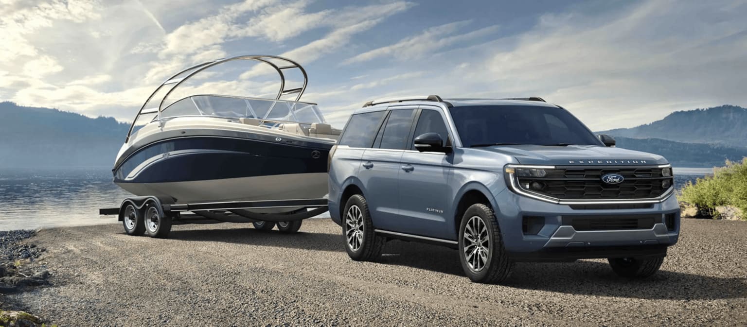 A large, rugged-looking SUV towing a sleek, modern-looking boat on a trailer, set against a scenic backdrop of mountains and cloudy skies.