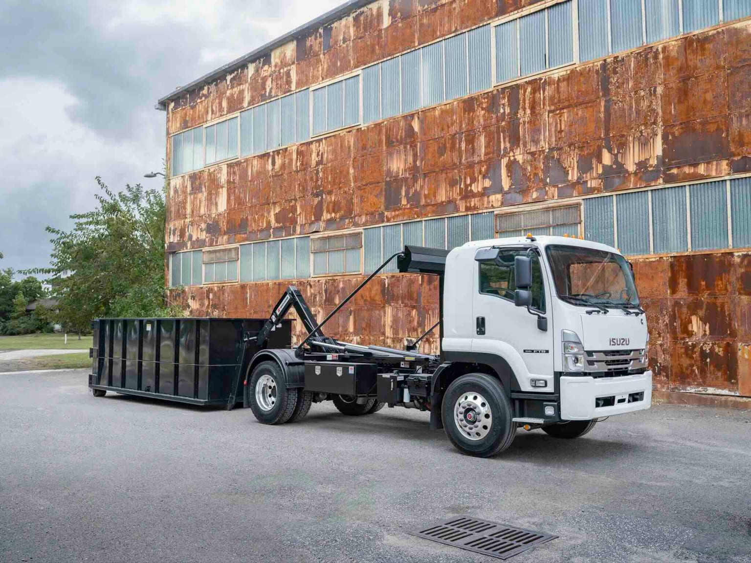 A white truck with a black dumpster trailer parked in front of a large, rusty industrial building.
