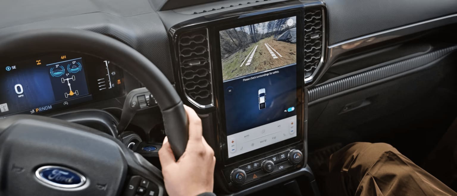 A hand grips the steering wheel of a vehicle, with a digital dashboard displaying speed and drive mode, and a large touchscreen showing a rearview camera feed of a dirt road.