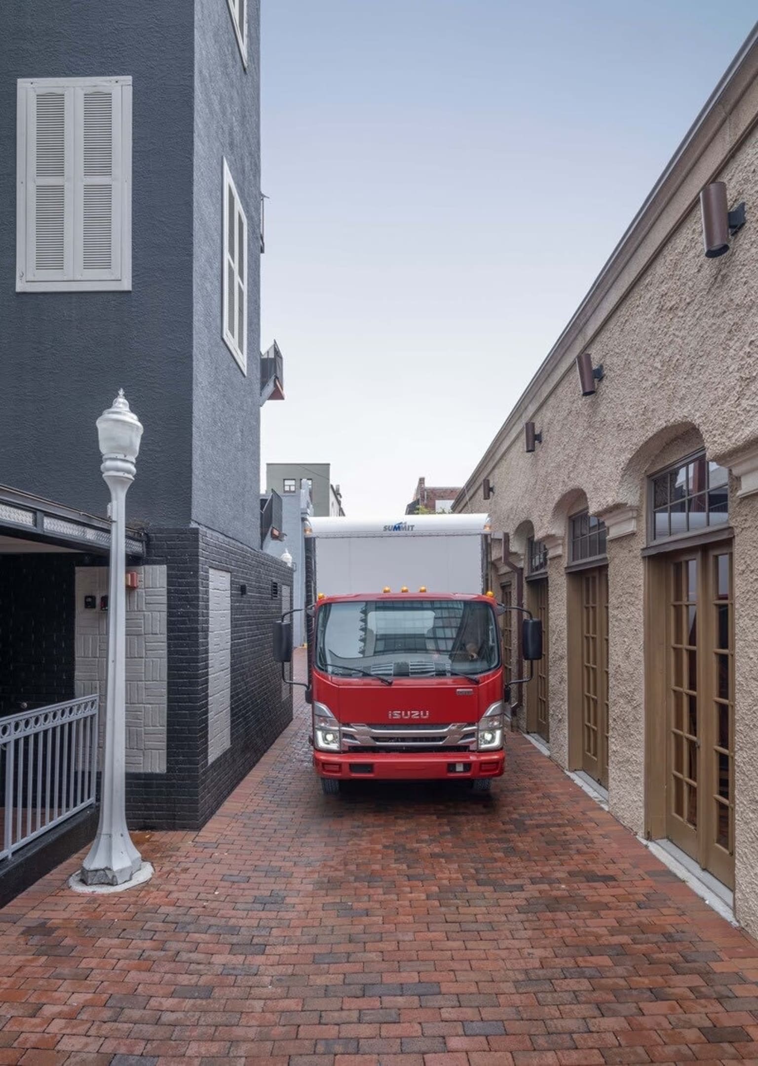 A red truck drives down a brick-paved alleyway between two buildings, one dark with white shutters and the other light-colored with arched doorways.