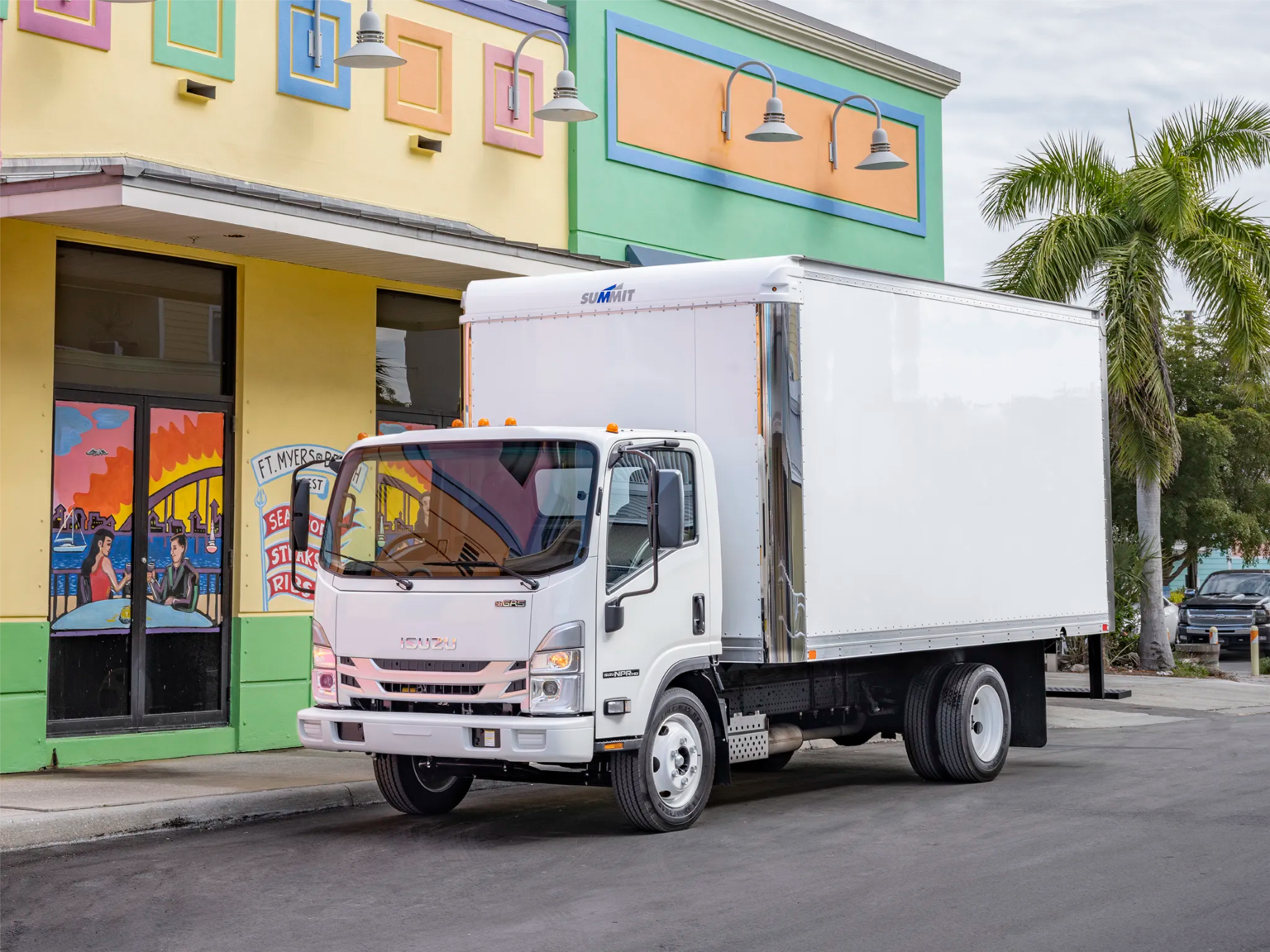 A white box truck parked on a street in front of a colorful building with palm trees in the background.