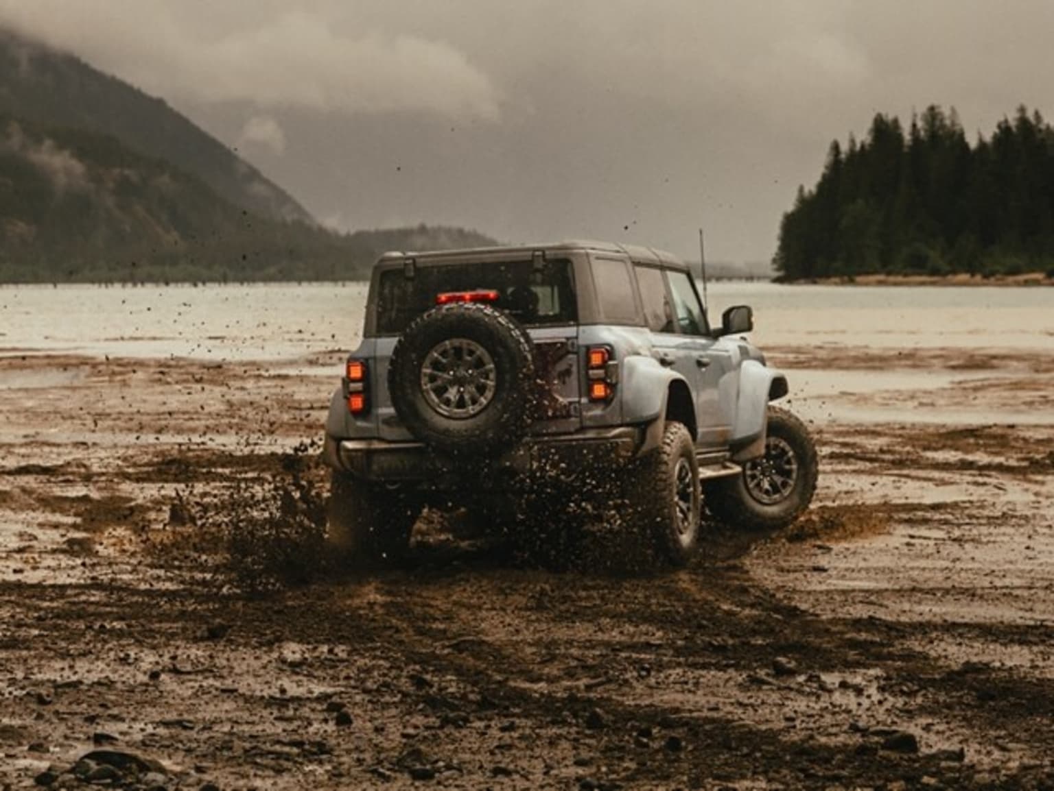 Off-road vehicle navigating muddy terrain with mountains and cloudy sky in the background.