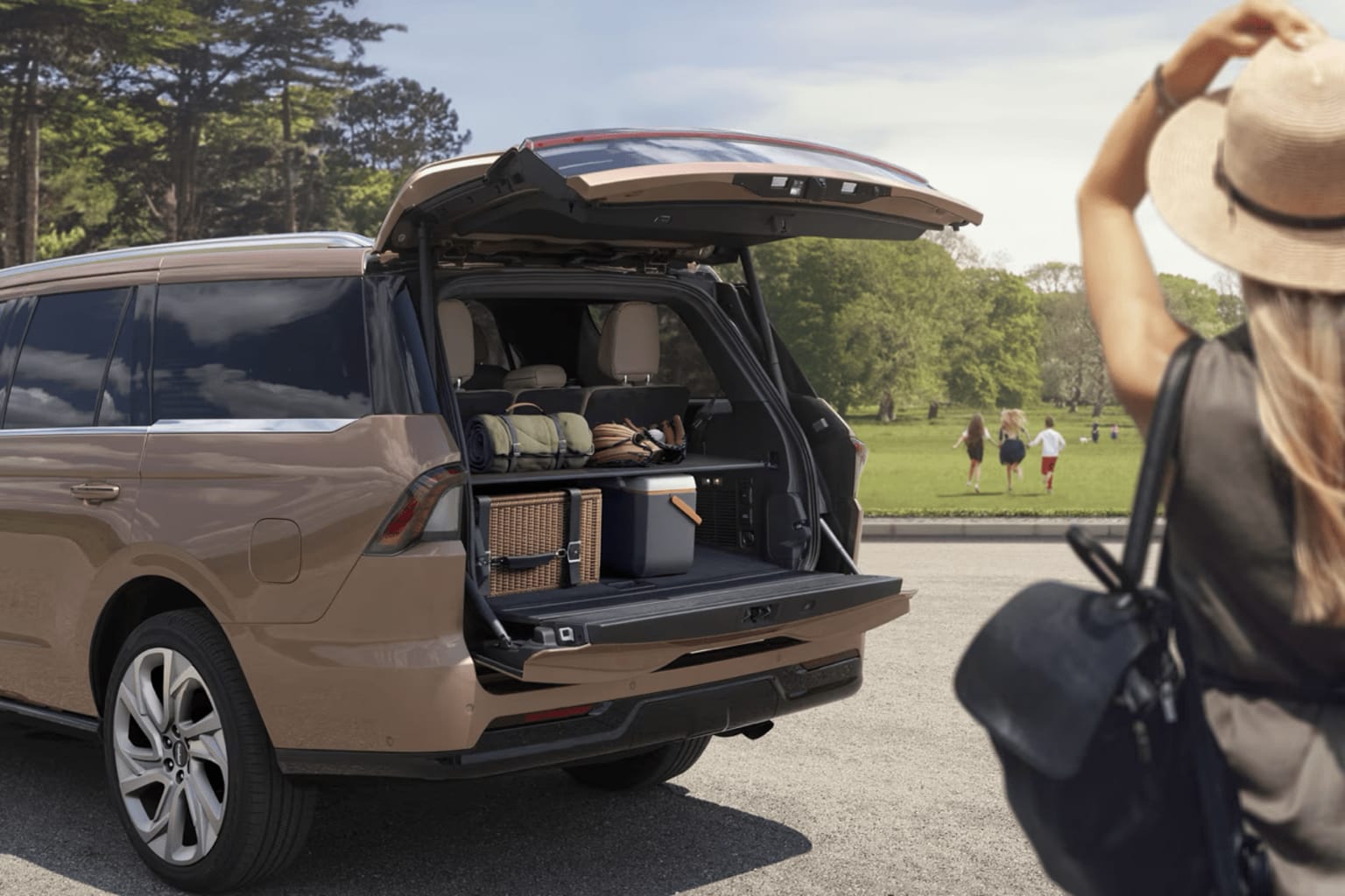 Beige SUV with an open trunk parked on a paved road surrounded by trees and a grassy field in the background.