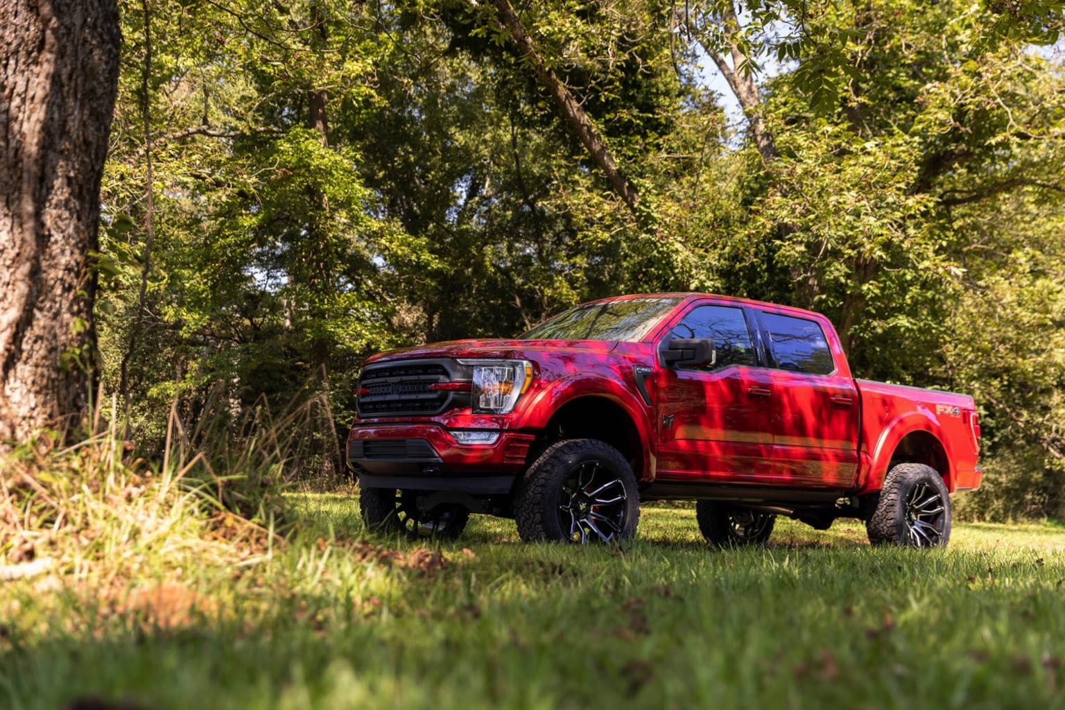 A red, lifted pickup truck sits in a grassy field surrounded by lush, green trees.