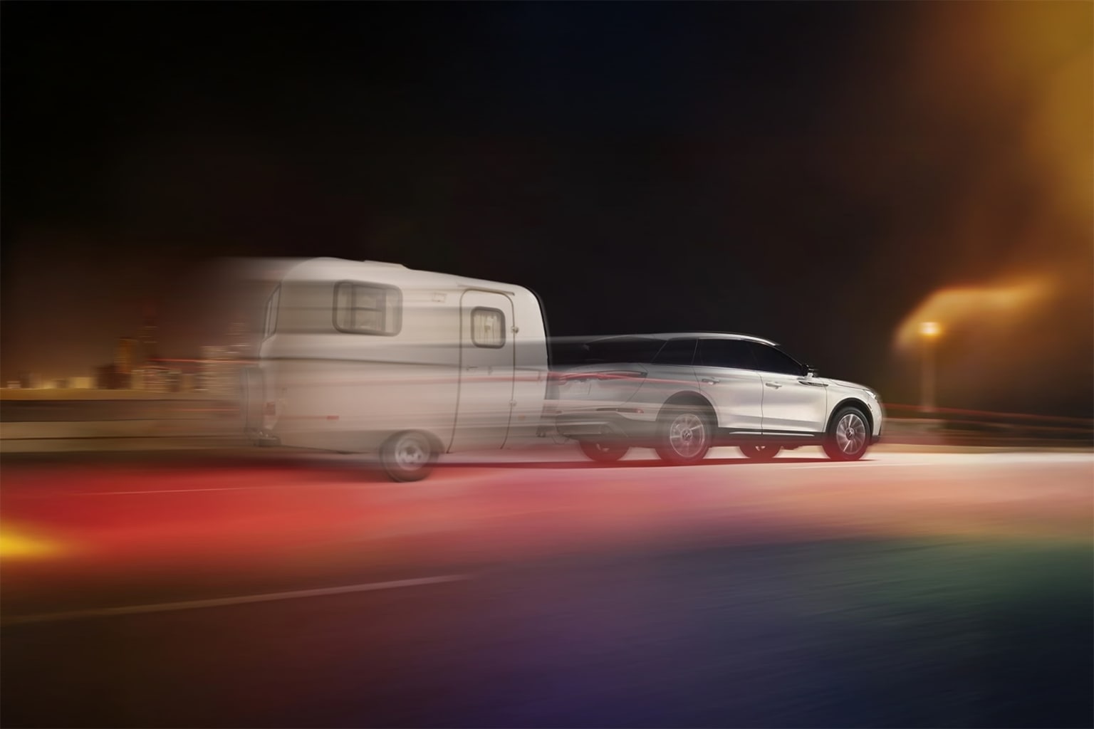 A white SUV towing a camper trailer on a dimly lit road with colorful lights in the background.