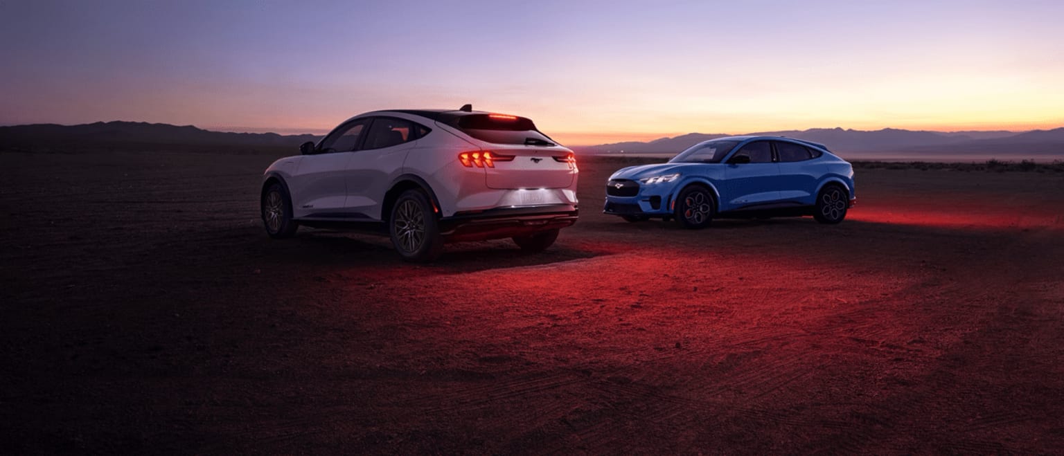 Two modern SUVs parked in a desert landscape at sunset, with their headlights and taillights illuminating the scene against the vibrant colors of the sky.