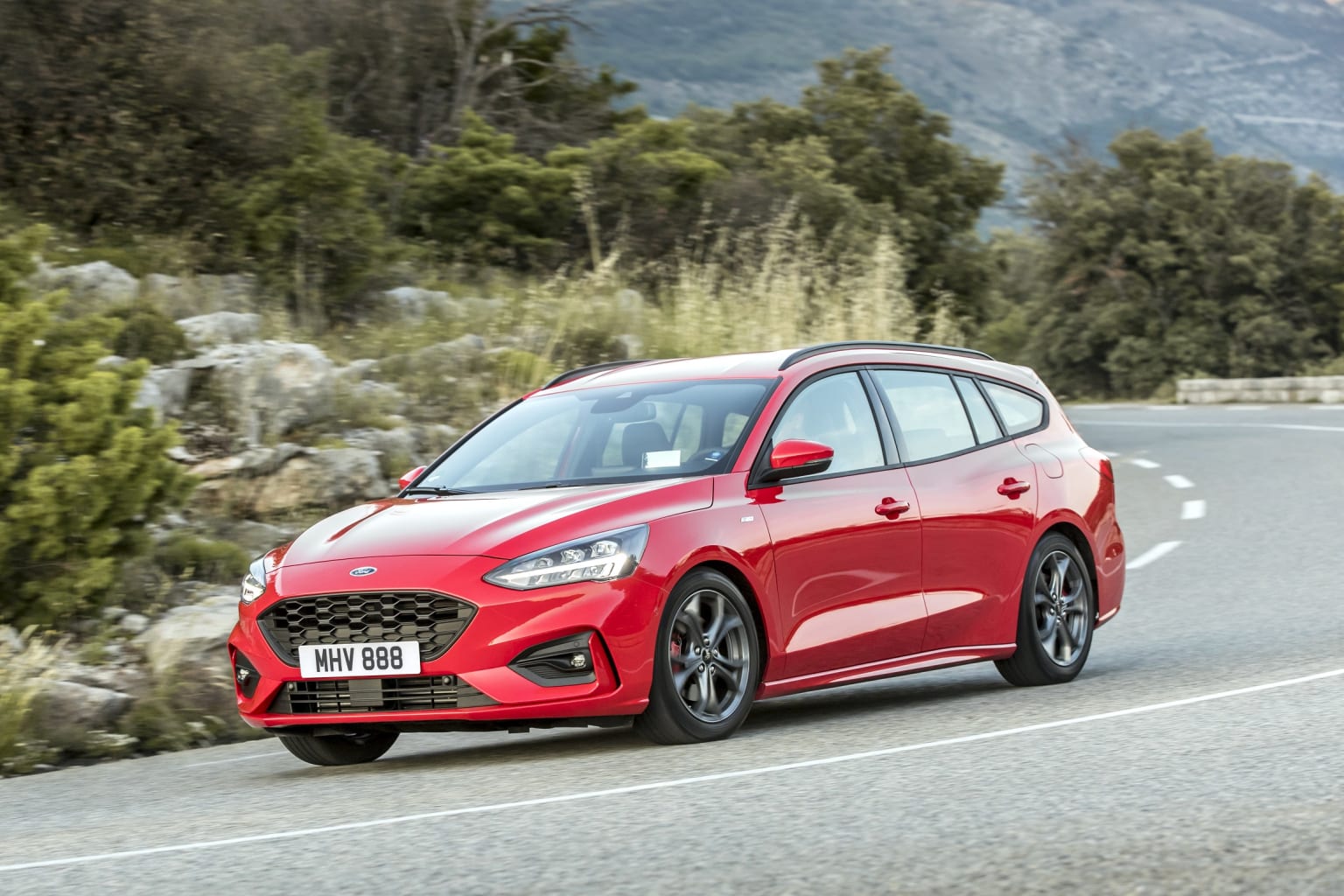 a red ford focus st driving down a road in front of a mountain range with trees and bushes on either side of the road