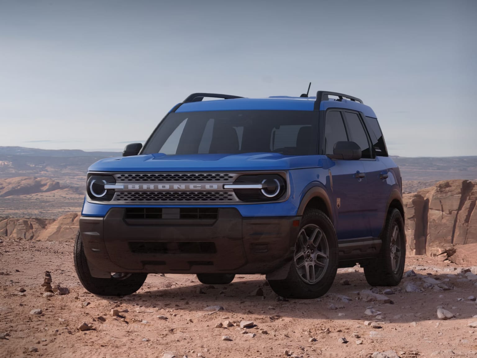 A blue compact SUV sits on a rocky desert plateau against a backdrop of red rock formations and a clear sky.