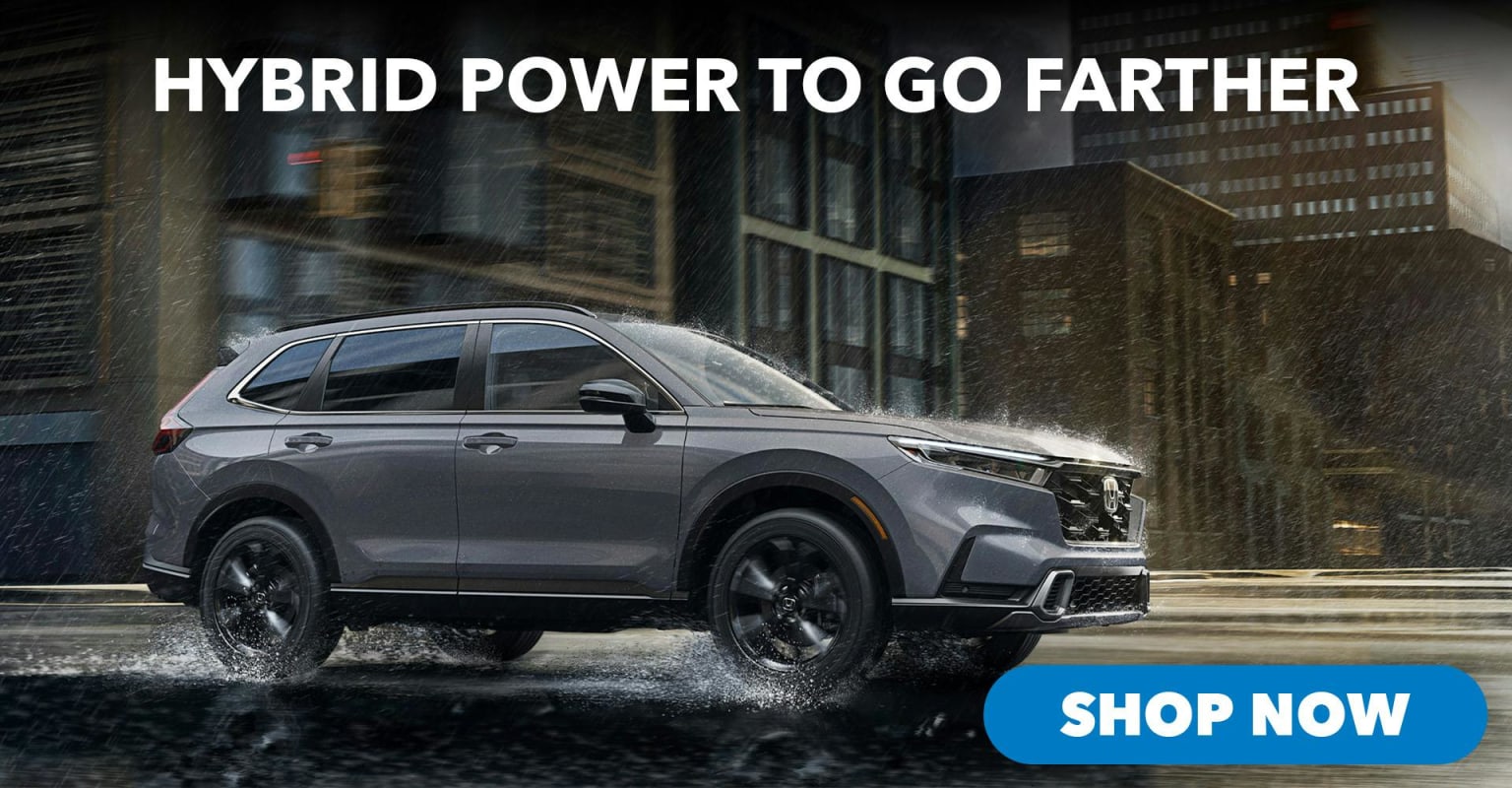 A gray hybrid SUV driving through a wet city street with tall buildings in the background, and a "SHOP NOW" button in the foreground.