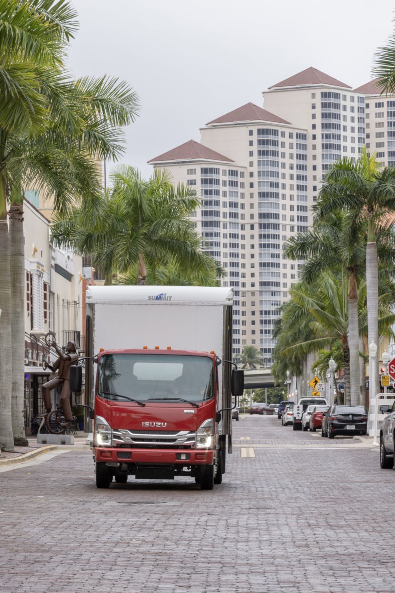A red truck drives down a cobblestone street lined with palm trees, with tall, modern buildings in the background.