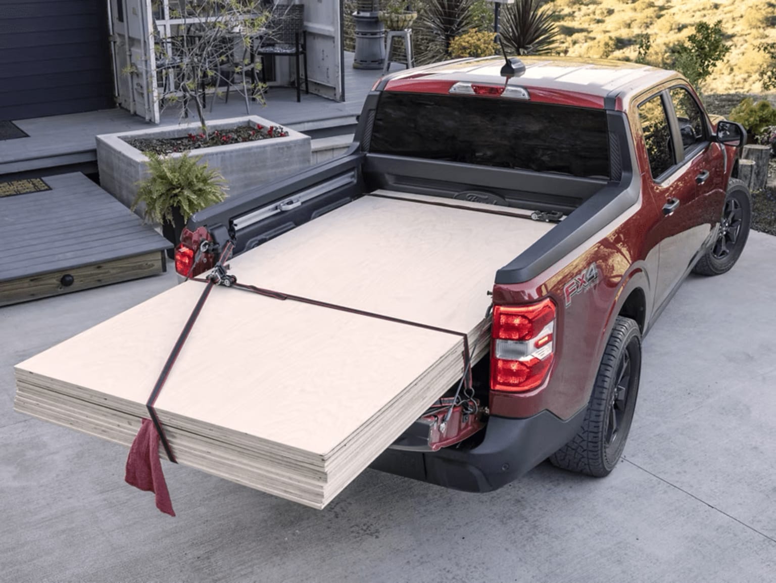 A red pickup truck with a cargo bed filled with large sheets of plywood or drywall, parked in front of a building with potted plants and a paved walkway.