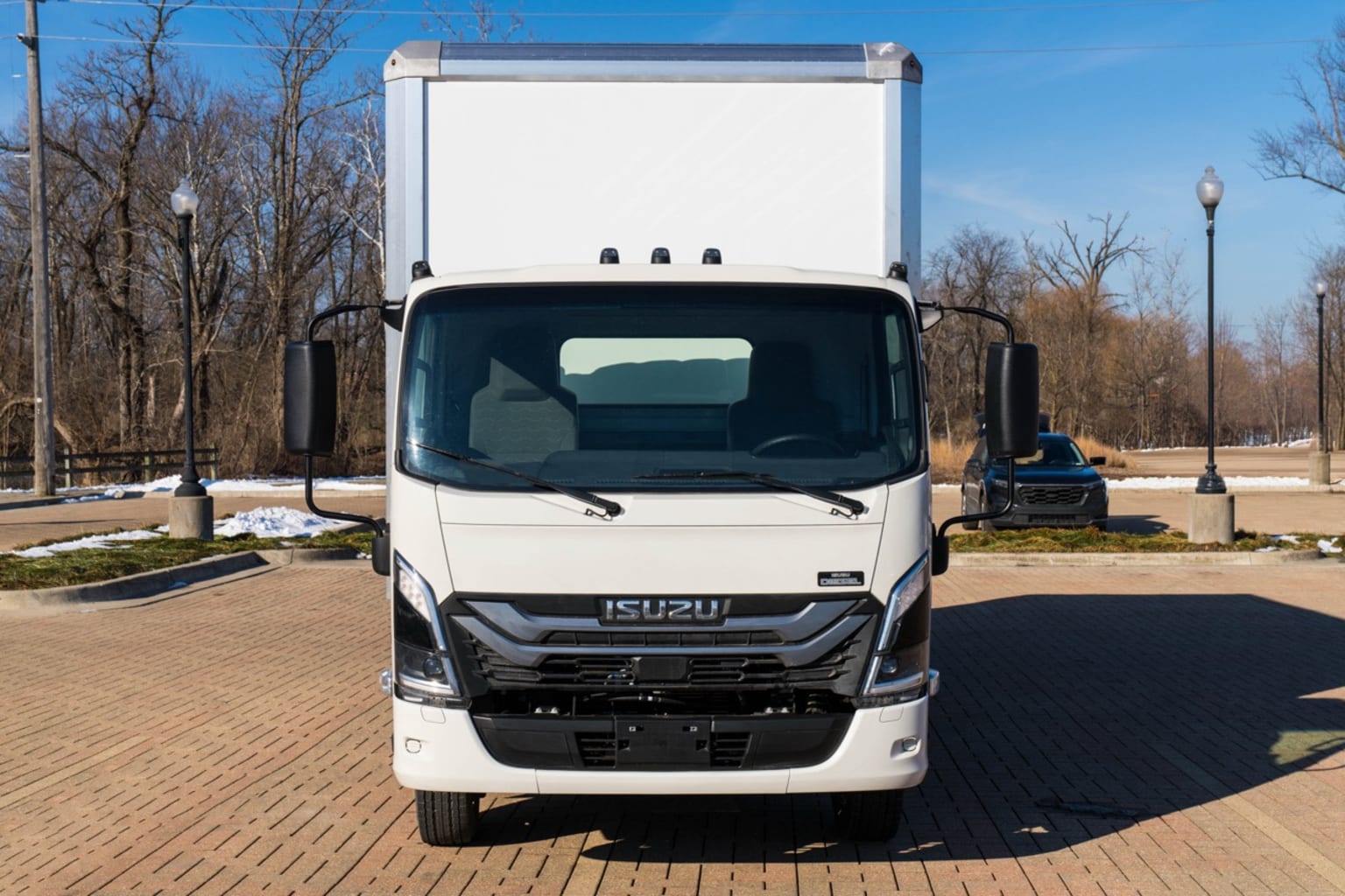 A white box truck is parked on a brick-paved lot with bare trees and a dark SUV in the background.
