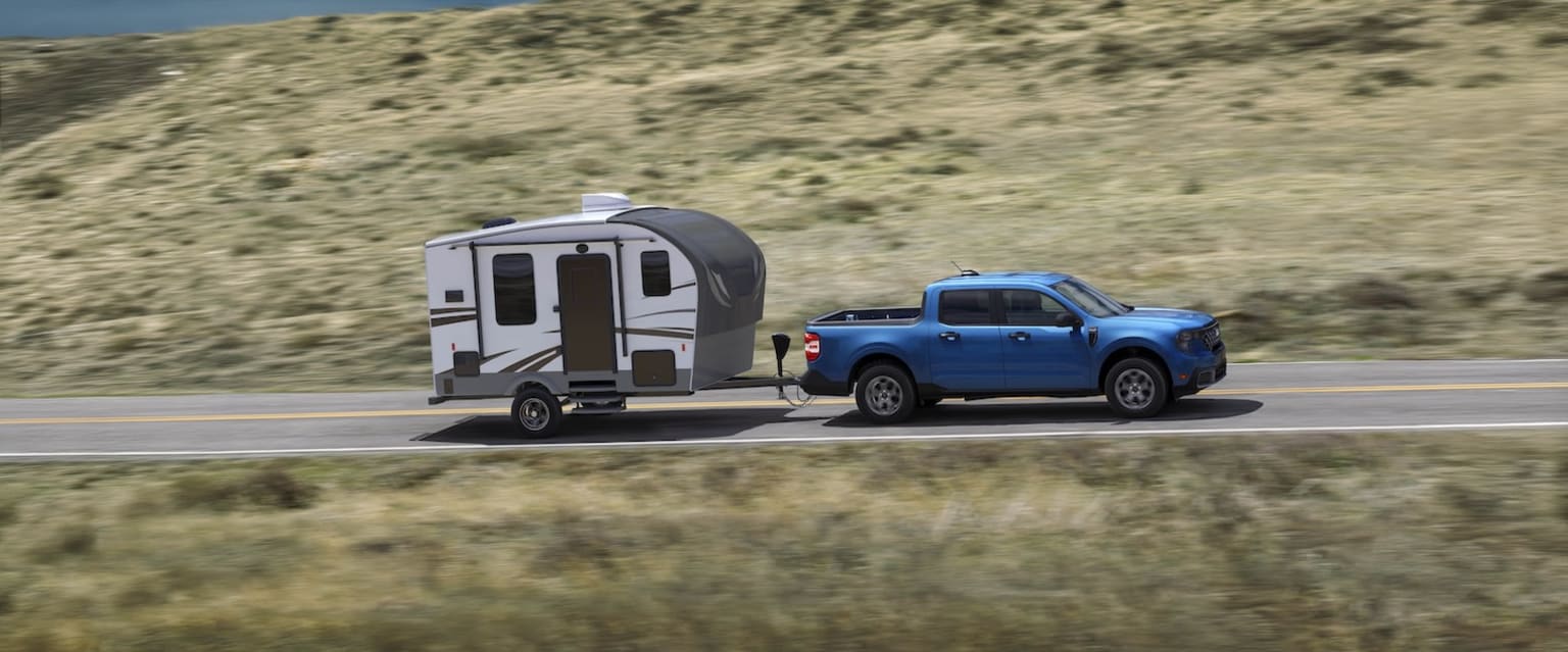 A blue pickup truck towing a small camper trailer travels along a winding road through a rugged, arid landscape.