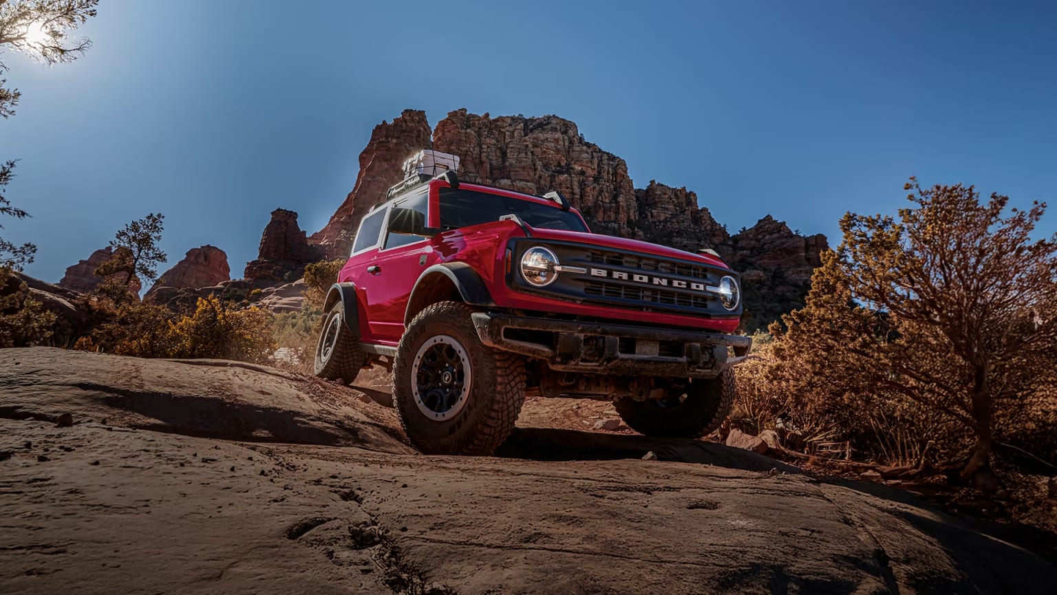 A red SUV drives on a rocky, desert trail with rugged mountains and a clear blue sky in the background.