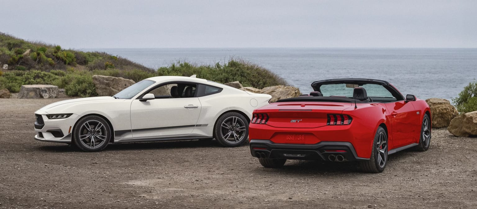 High-performance sports cars, a white coupe and a red convertible, parked on a dirt road overlooking a scenic coastal landscape with a body of water and vegetation in the background