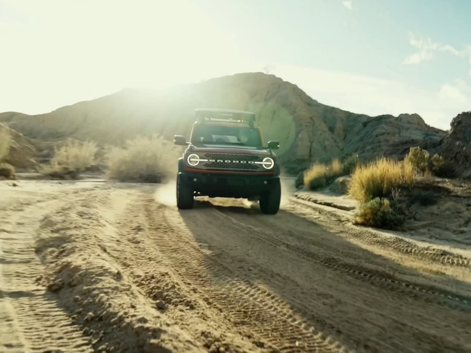 A red SUV kicks up dust on a dirt road in a desert landscape with mountains in the background.