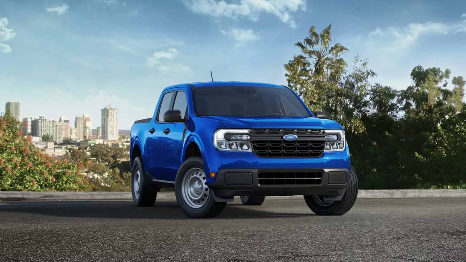 A blue pickup truck is parked on an asphalt road with a city skyline and trees in the background under a cloudy sky.