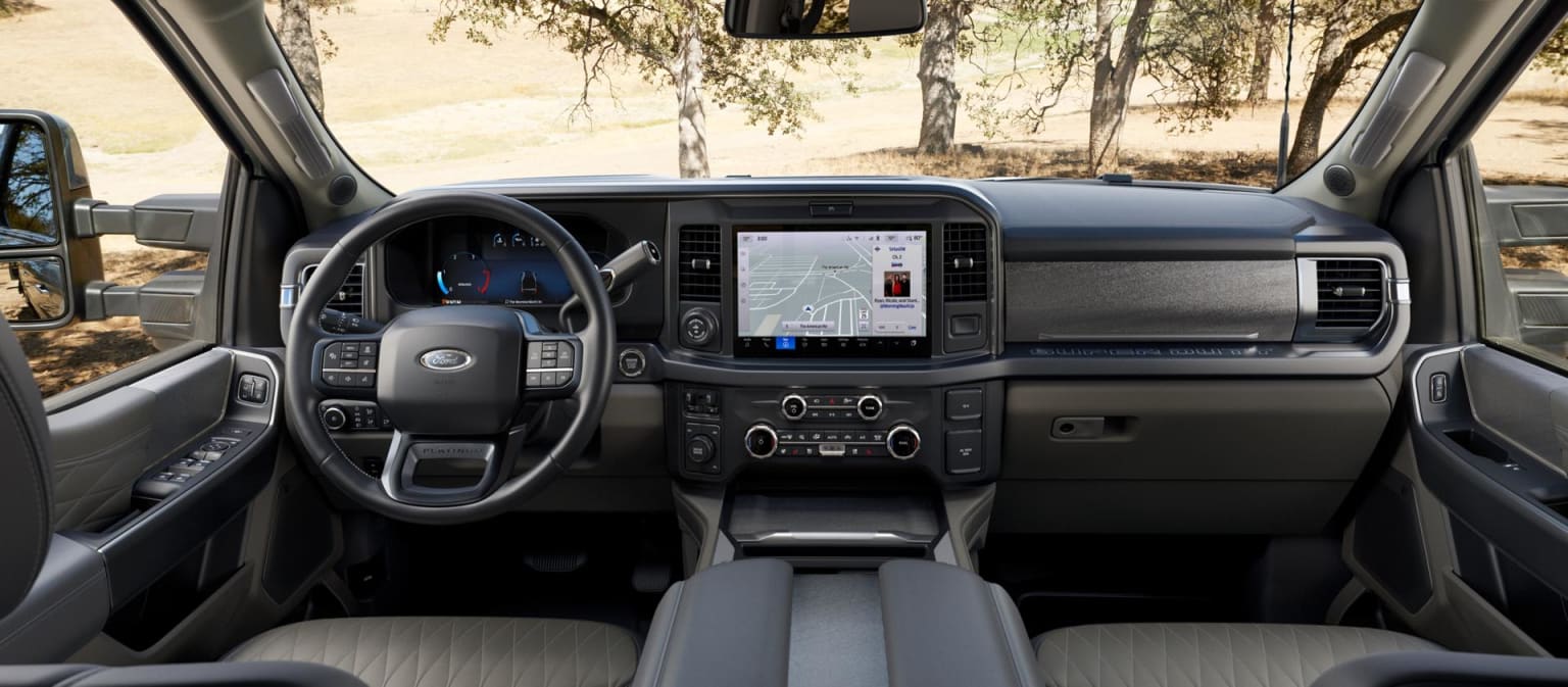 Interior view of a vehicle dashboard with a touchscreen display, surrounded by a wooded area visible through the windshield.