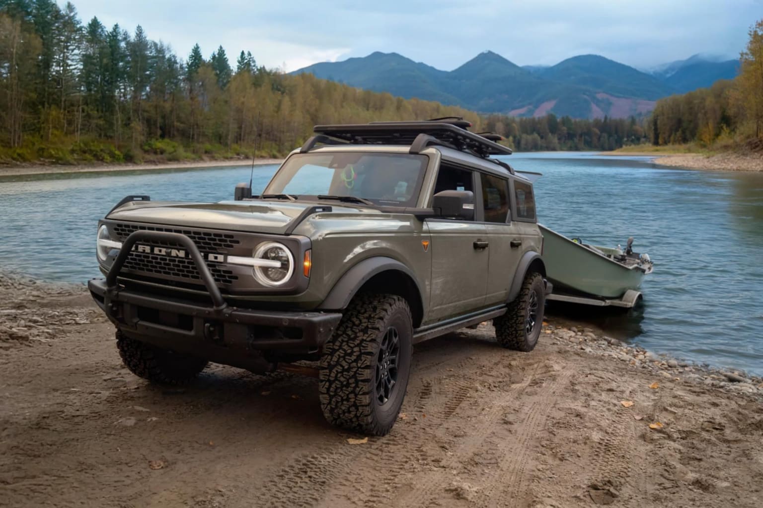 An off-road vehicle with a boat trailer parked on a sandy riverbank, with a calm river and forested mountains in the background.