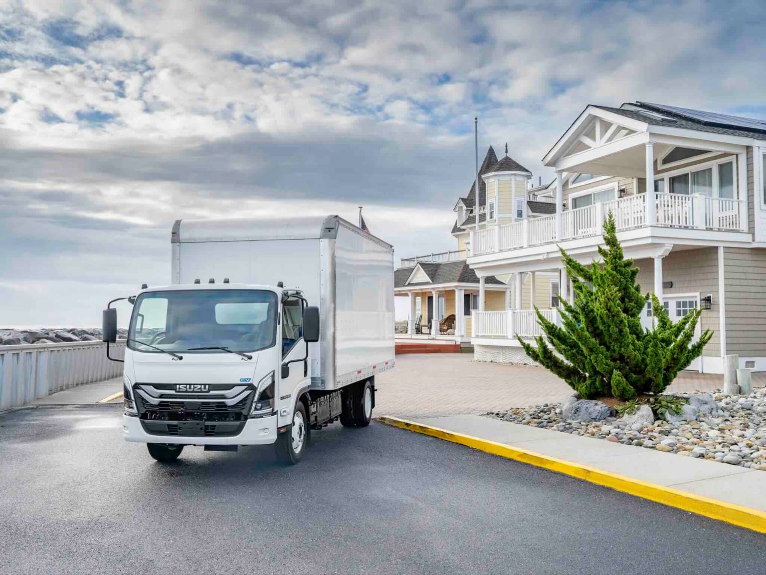A white box truck parked on a wet road in front of a large, ornate house with a cloudy sky overhead.