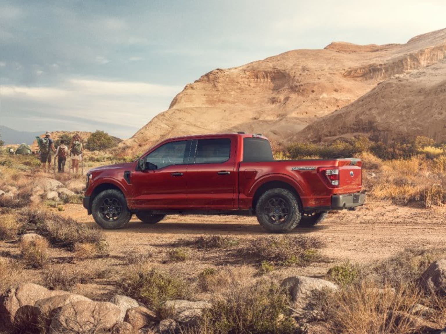a red pick up truck parked on a dirt road in the desert with a group of people in the background