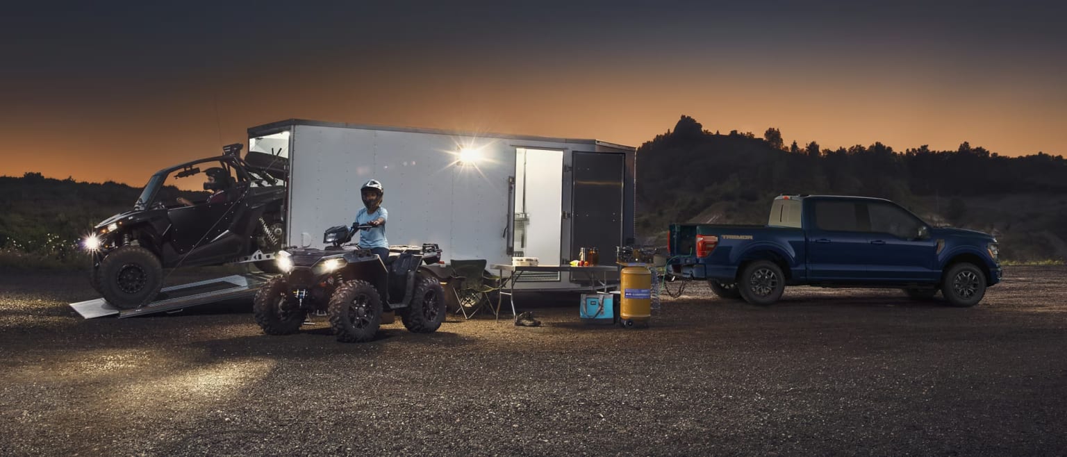 A nighttime outdoor scene with a large truck, a smaller truck, and a trailer set up in a remote, forested area against a backdrop of a dramatic sunset sky.