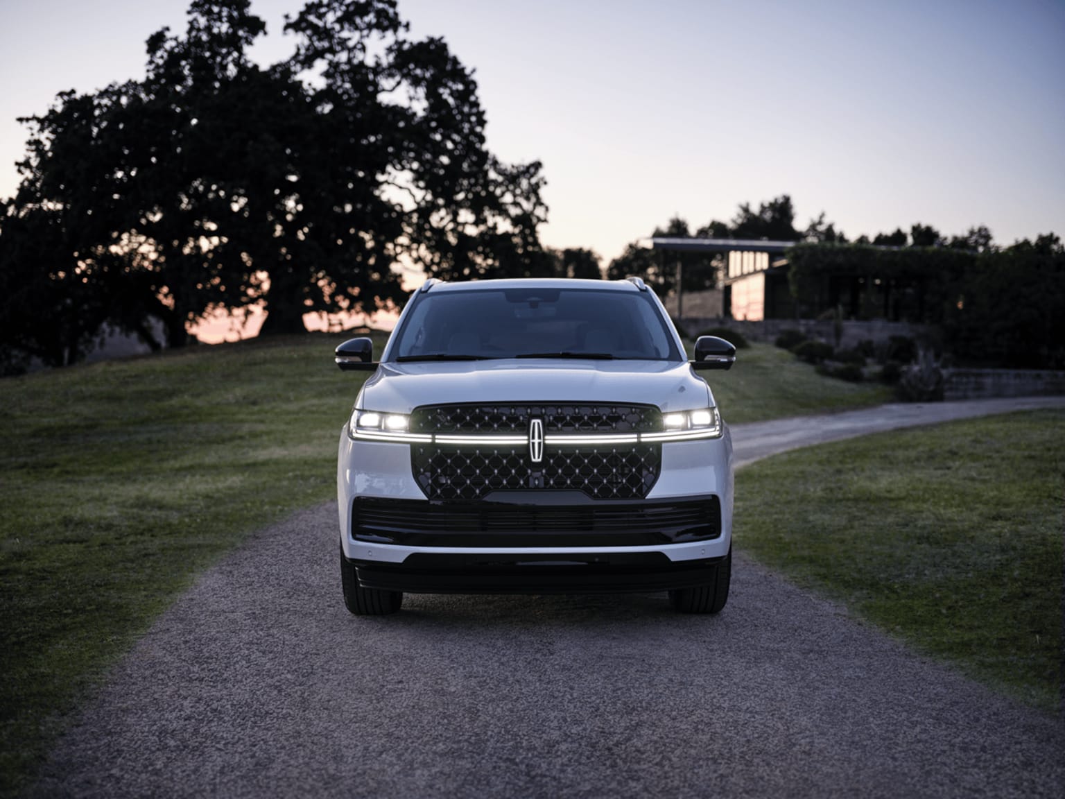 A large, silver SUV on a gravel driveway surrounded by lush greenery with a sunset sky in the background.