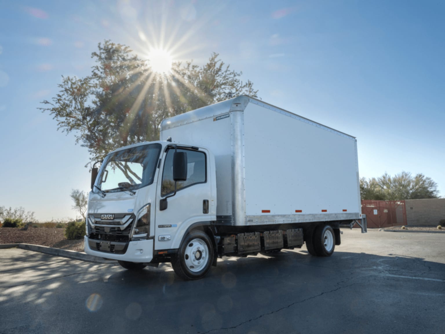 A white box truck parked on a paved lot under a sunny sky with a tree in the background.
