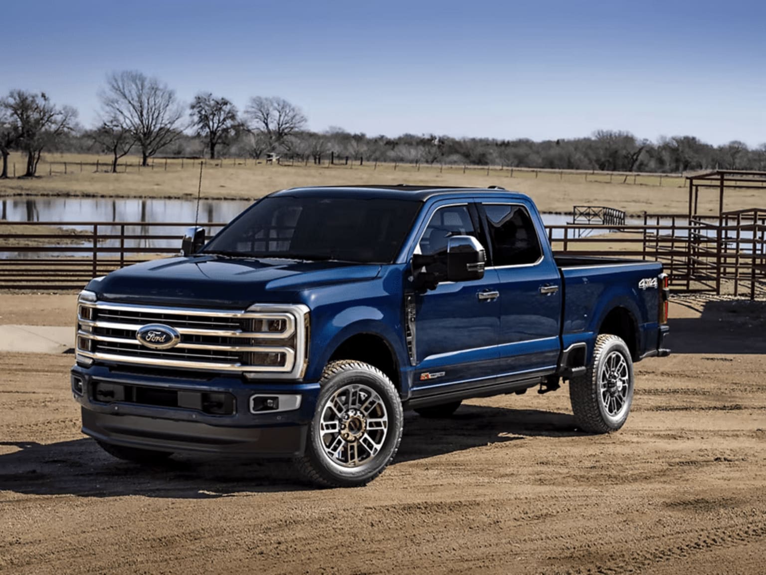 A blue pickup truck is parked on a dirt road in a rural setting, with a fenced field and trees visible in the background.