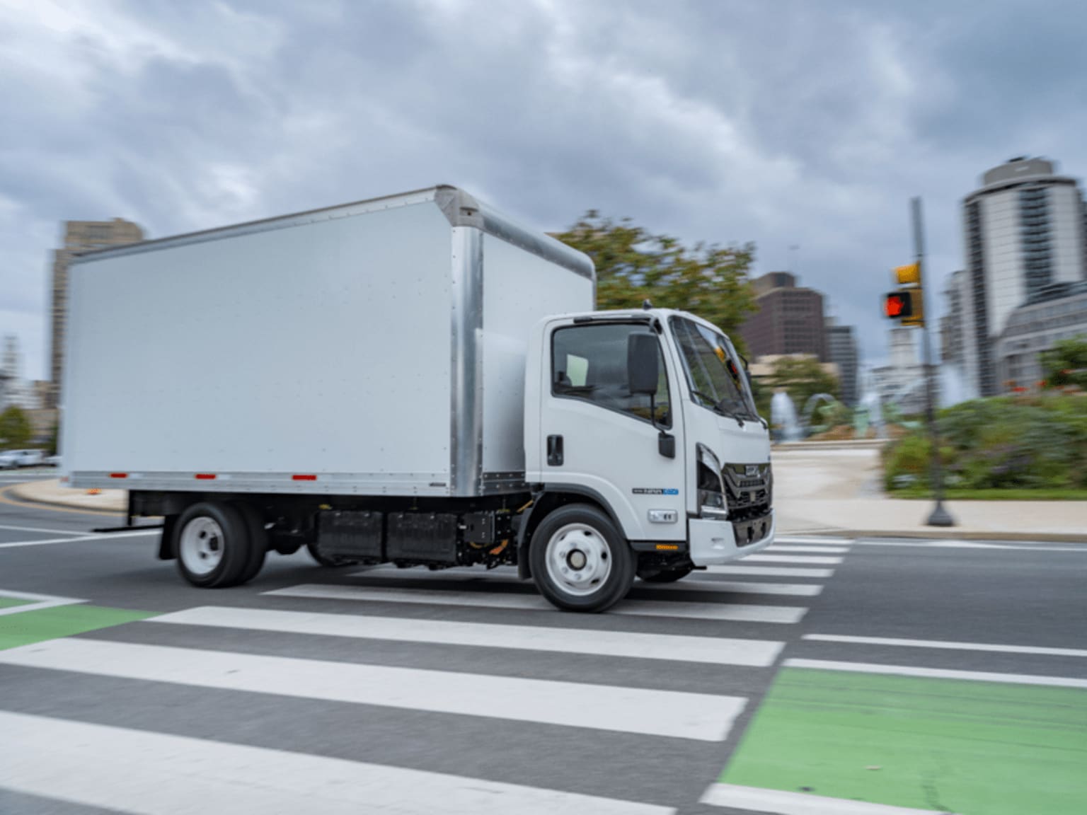 A white box truck crossing a city crosswalk with tall buildings and cloudy sky.