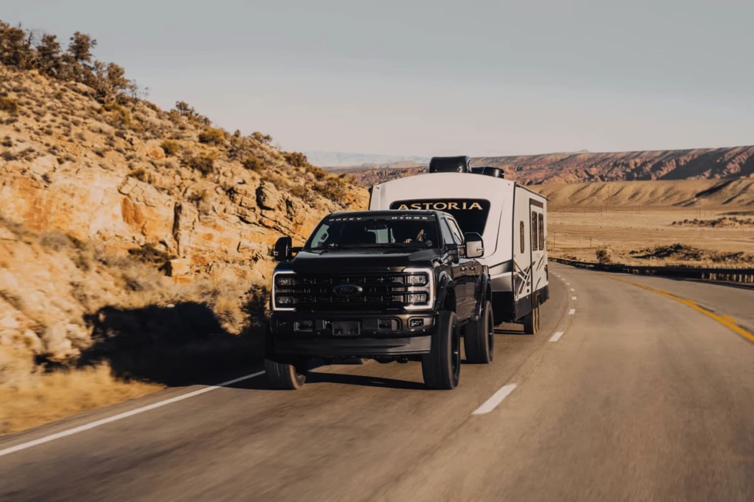 A large black pickup truck towing a white recreational vehicle travels down a winding road through a rugged, mountainous desert landscape.