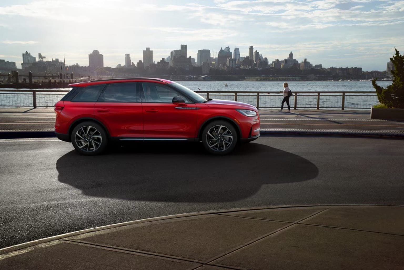 A bright red SUV parked on a waterfront promenade with a stunning cityscape of skyscrapers in the background across the water.
