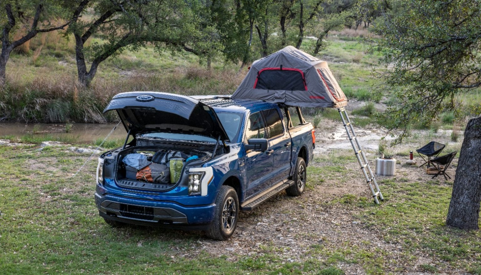 a truck with a tent on top of it parked next to a tree and a river in the background