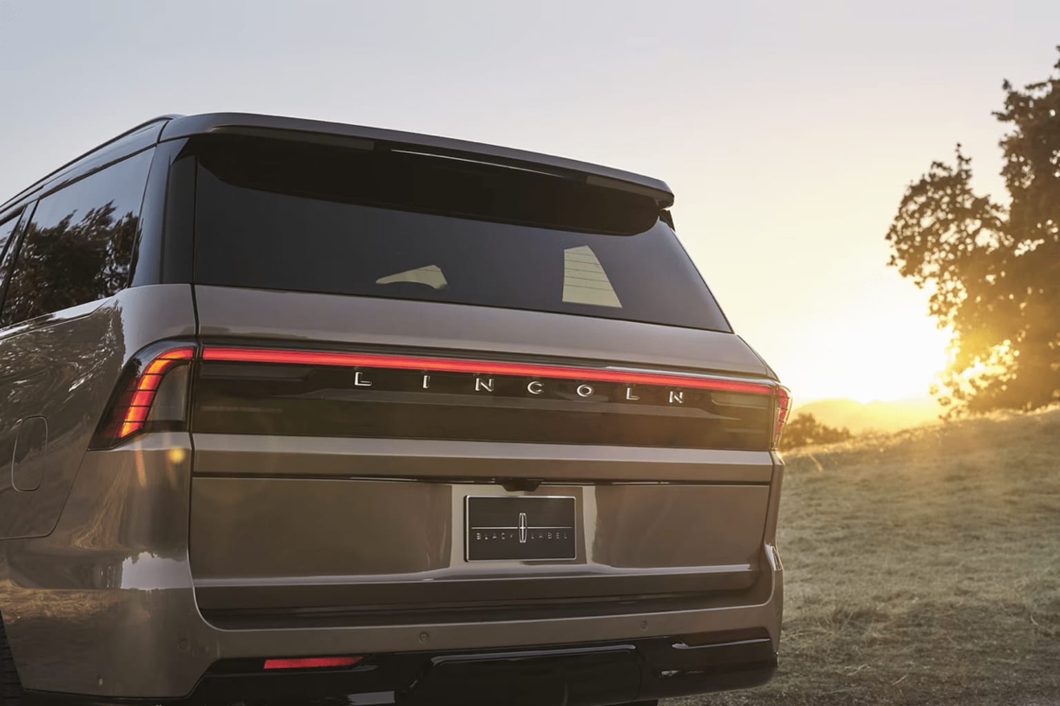 A large, dark-colored SUV is parked in a grassy field, with the sun setting in the background, casting a warm glow over the scene.