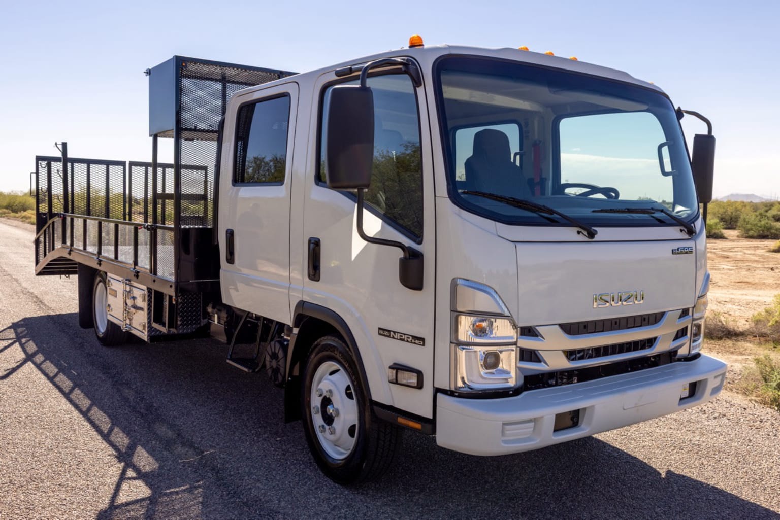 A white flatbed truck with a cage in the back on a paved road with a desert landscape in the background.
