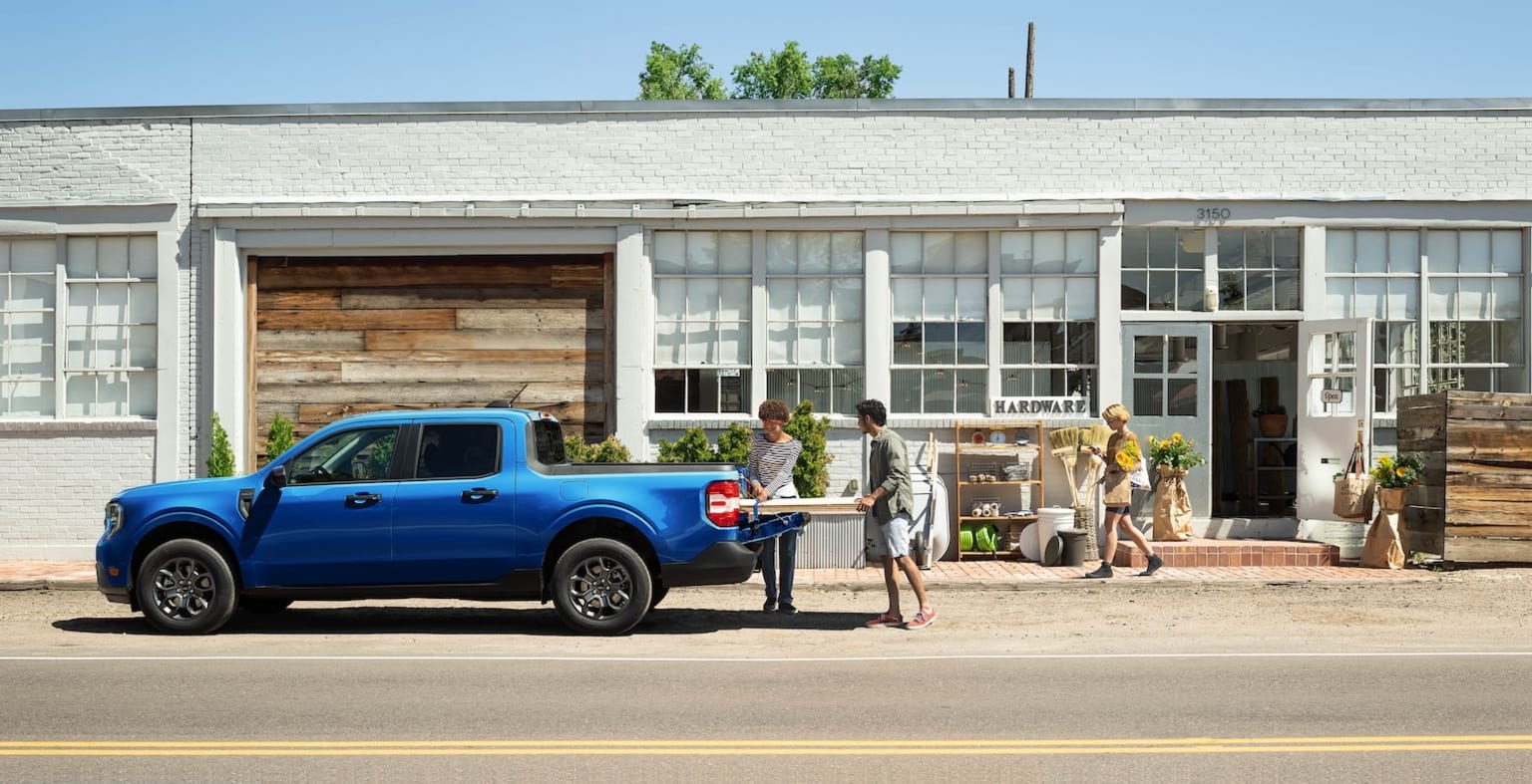 A blue pickup truck parked in front of a rustic-style building with large windows, surrounded by people standing outside the building.