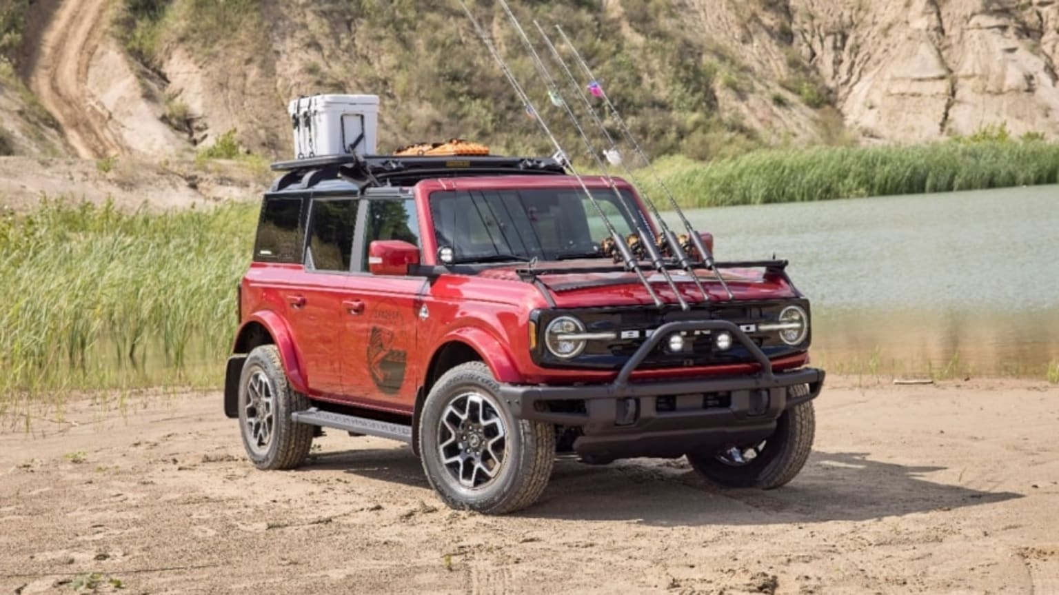 a red truck parked on a dirt road next to a body of water and a mountain range in the background