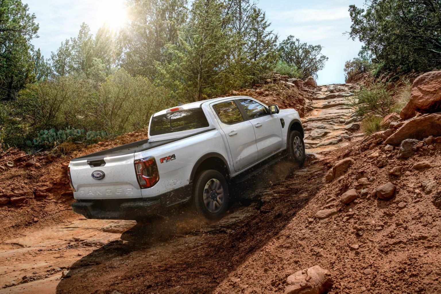 A white truck navigates a rocky, off-road trail with trees and red rock formations in the background.